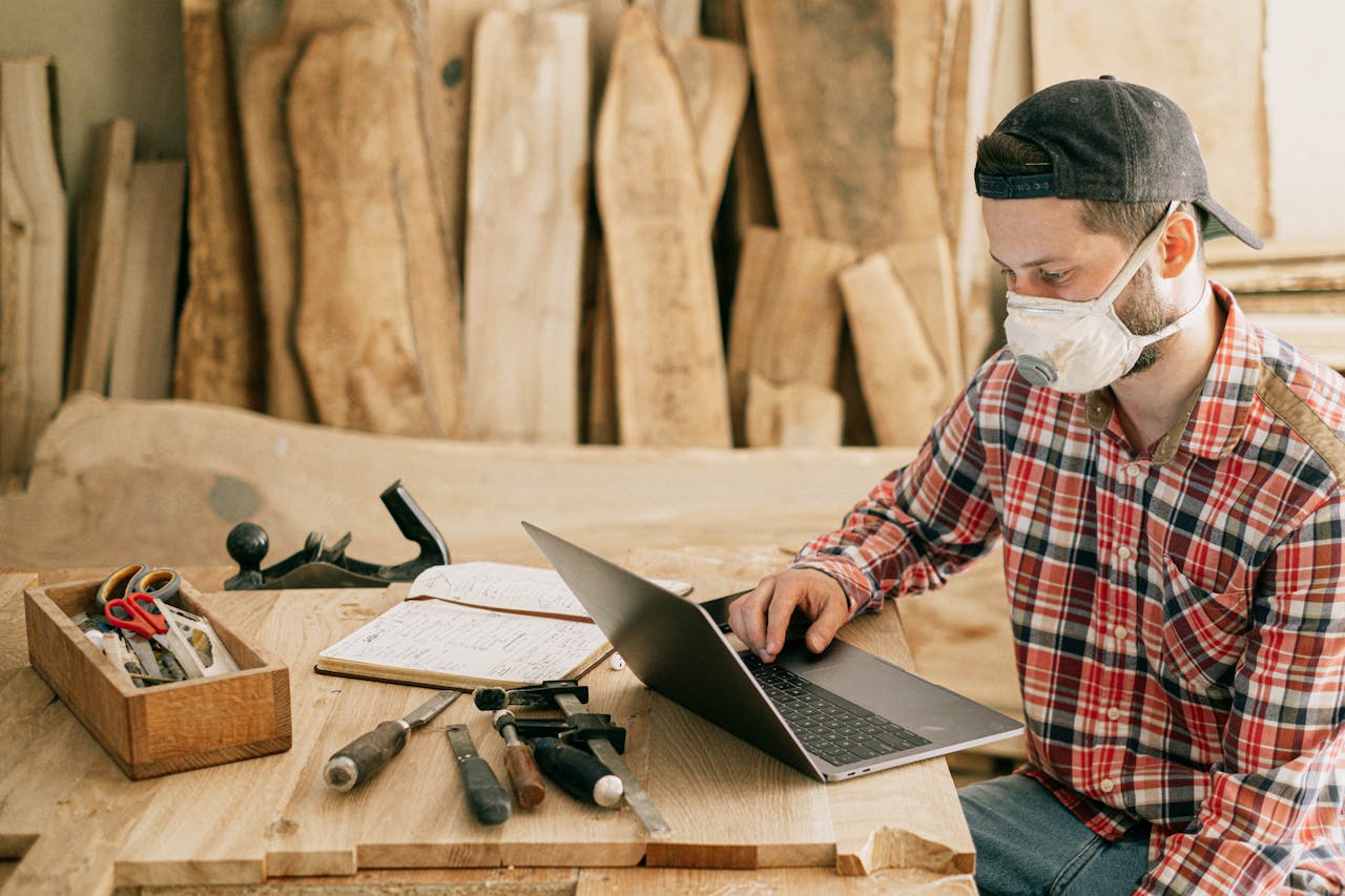 Craftsman working on wood with tools and laptop in workshop.