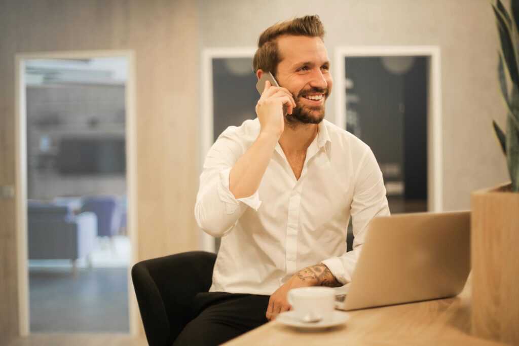 Smiling man talking on phone while working at laptop with coffee.