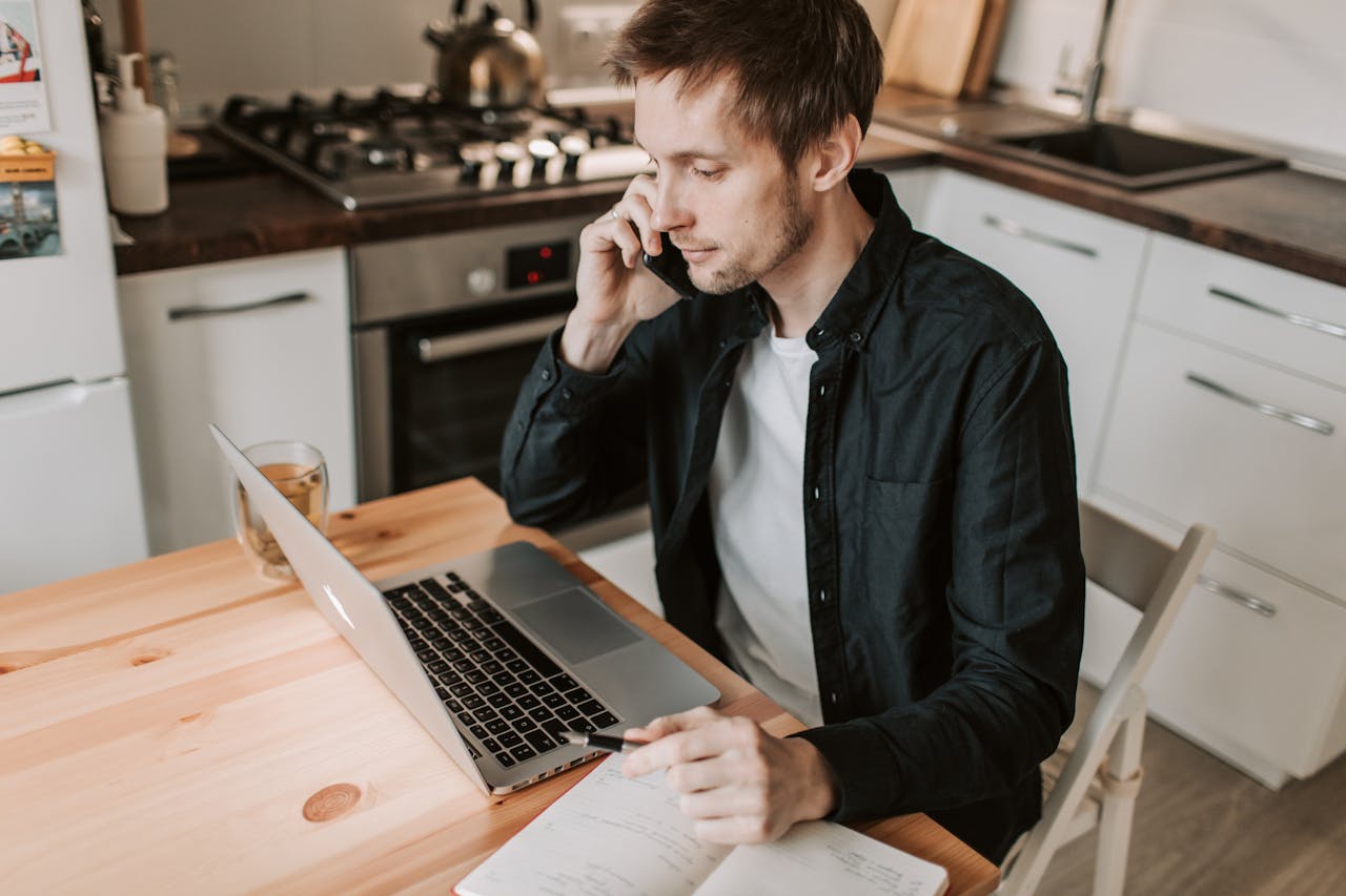 Man working on laptop while talking on phone in kitchen.