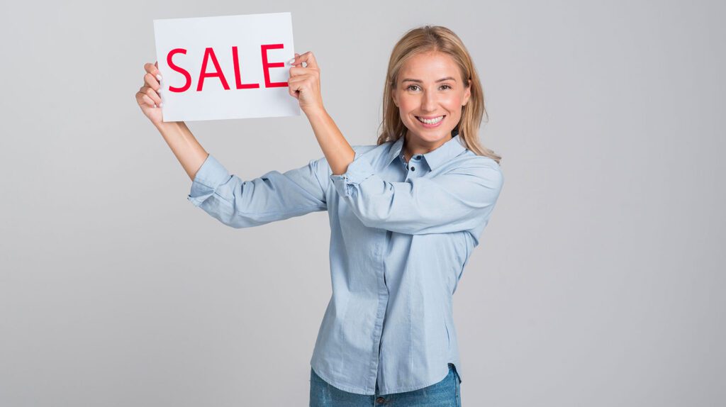 Smiling woman holding a SALE sign against a gray background.