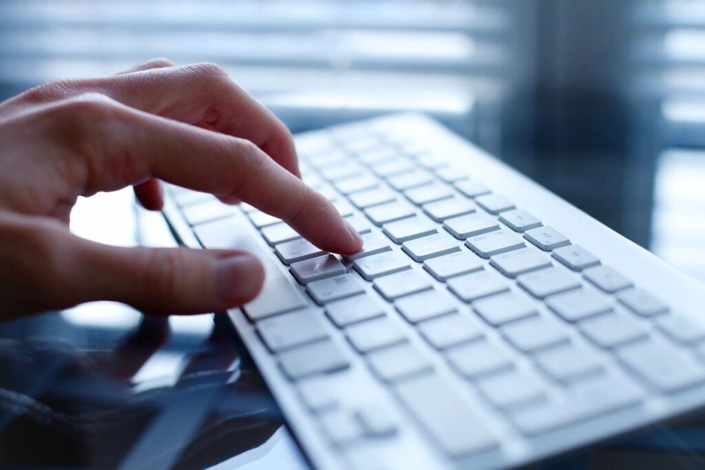 Person typing on a white keyboard near a window with blinds.