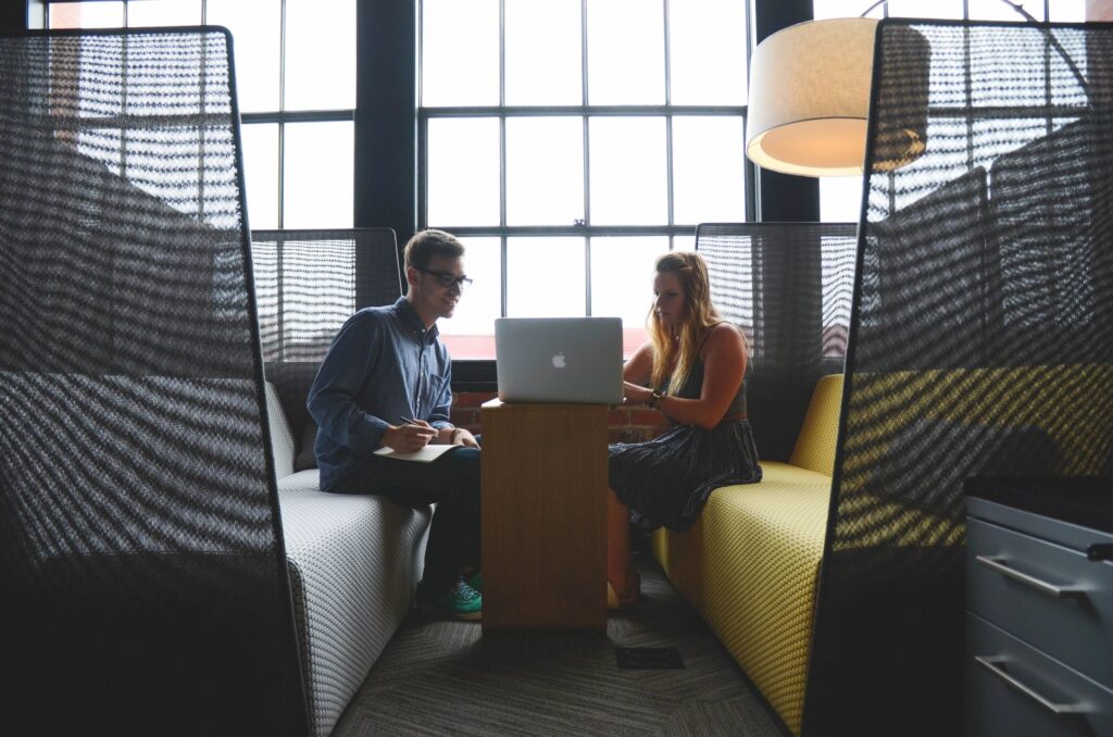 Two colleagues discussing work in a modern office booth with a laptop.
