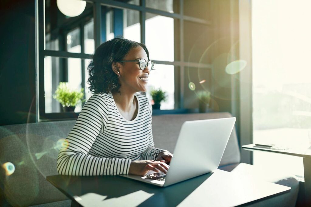 A woman working on a laptop near a window with natural light.