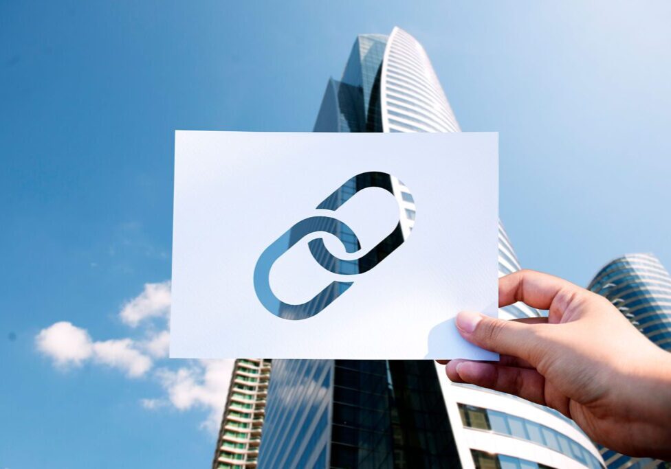 Hand holding a paper with a chain link icon against skyscrapers and blue sky.