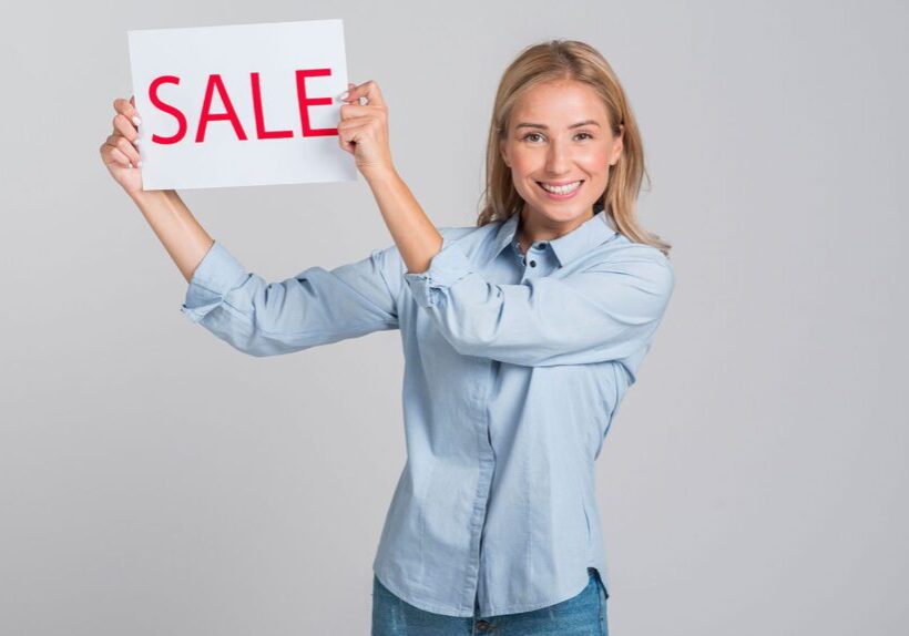 Smiling woman holding a SALE sign against a gray background.