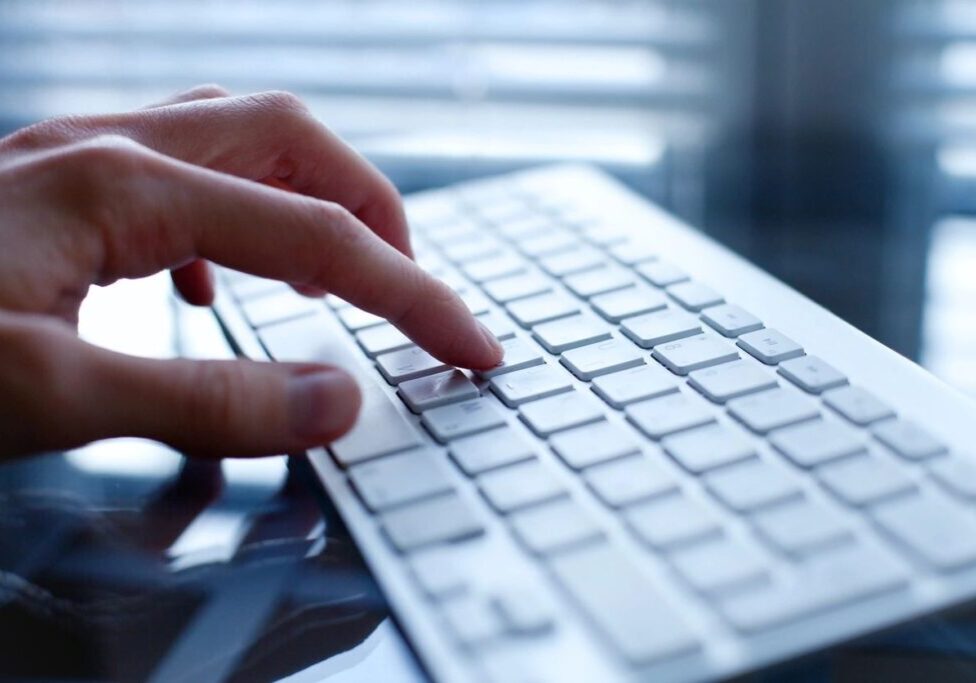 Person typing on a white keyboard near a window with blinds.