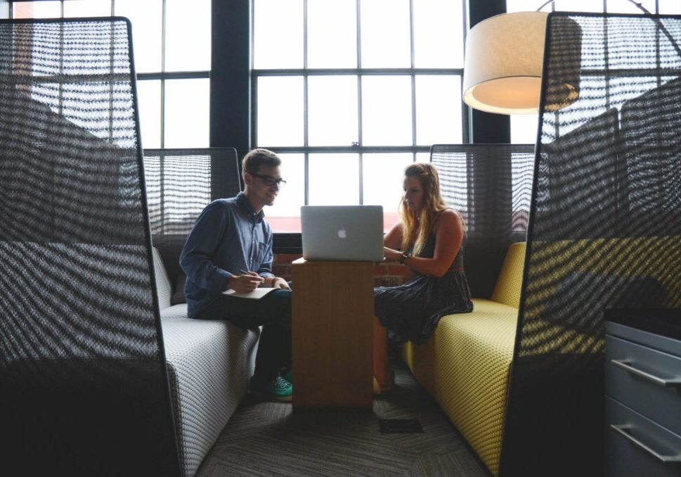 Two colleagues discussing work in a modern office booth with a laptop.